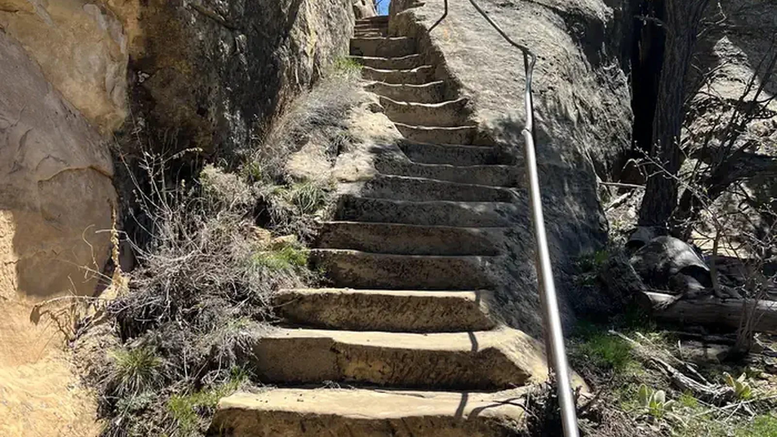 Steep stone steps descend through a narrow crack in a rock cliff; a metal railing parallels the steps and a blue sky is above.