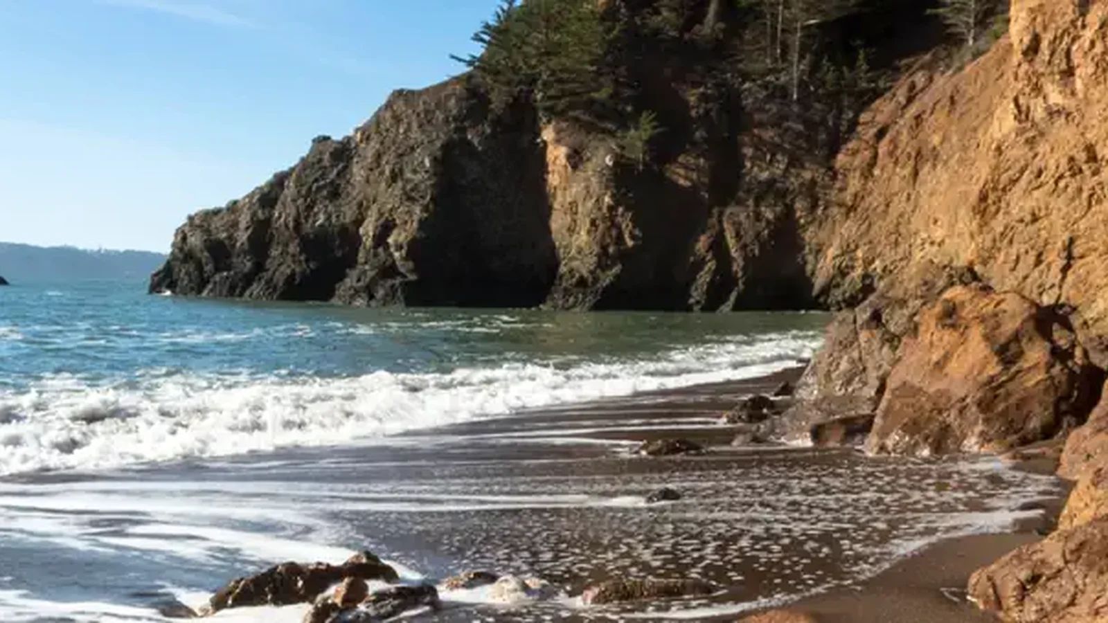 The beach at Kirby Cove, with dramatic cliffs, dark volcanic sand, and frothy water.