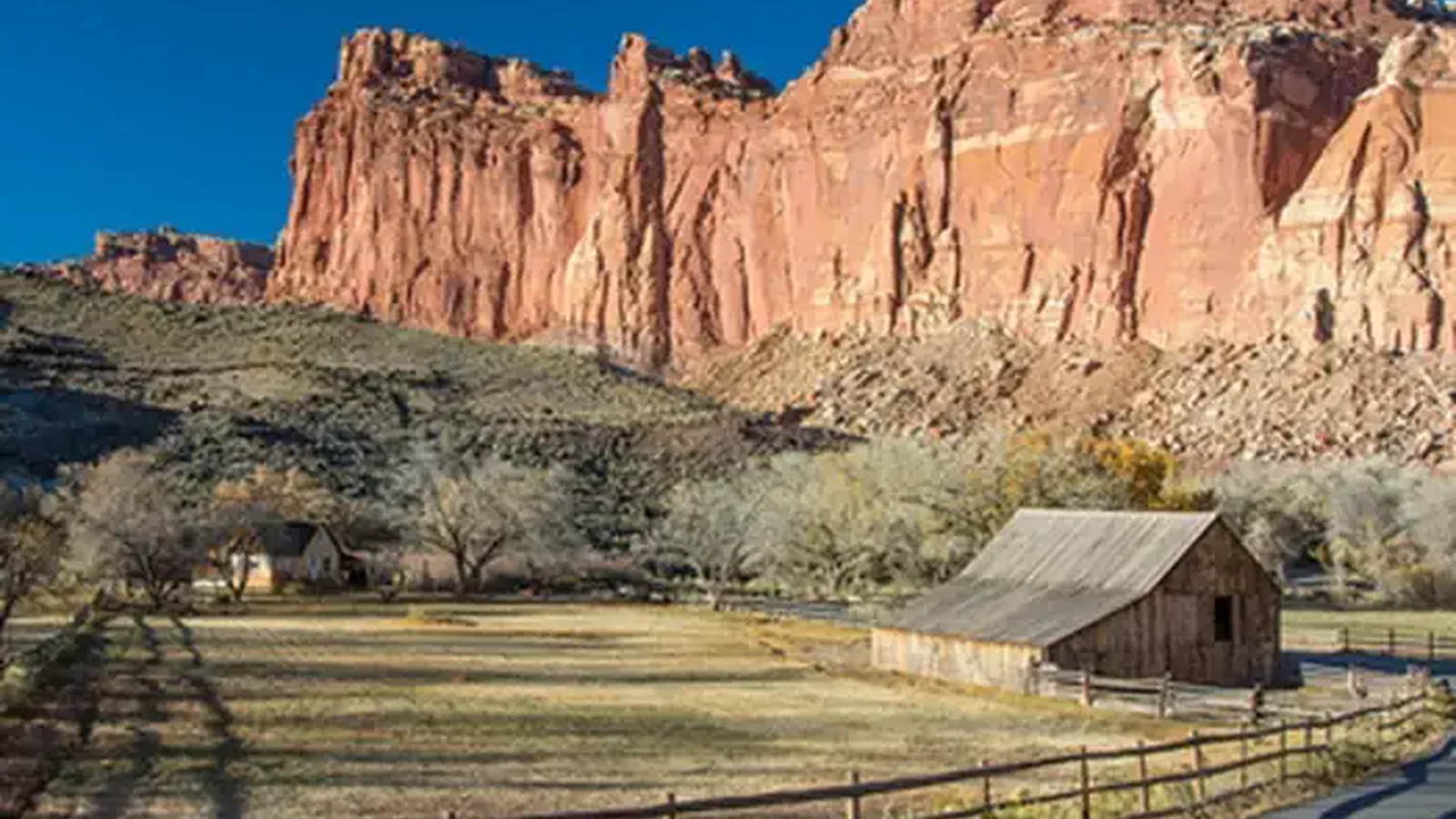 A brown barn is in a grassy field. A fence separates the field from the road. Behind the barn are leafless trees and orange-red rock rises above the landscape. The sky is a rich, cloudless blue.