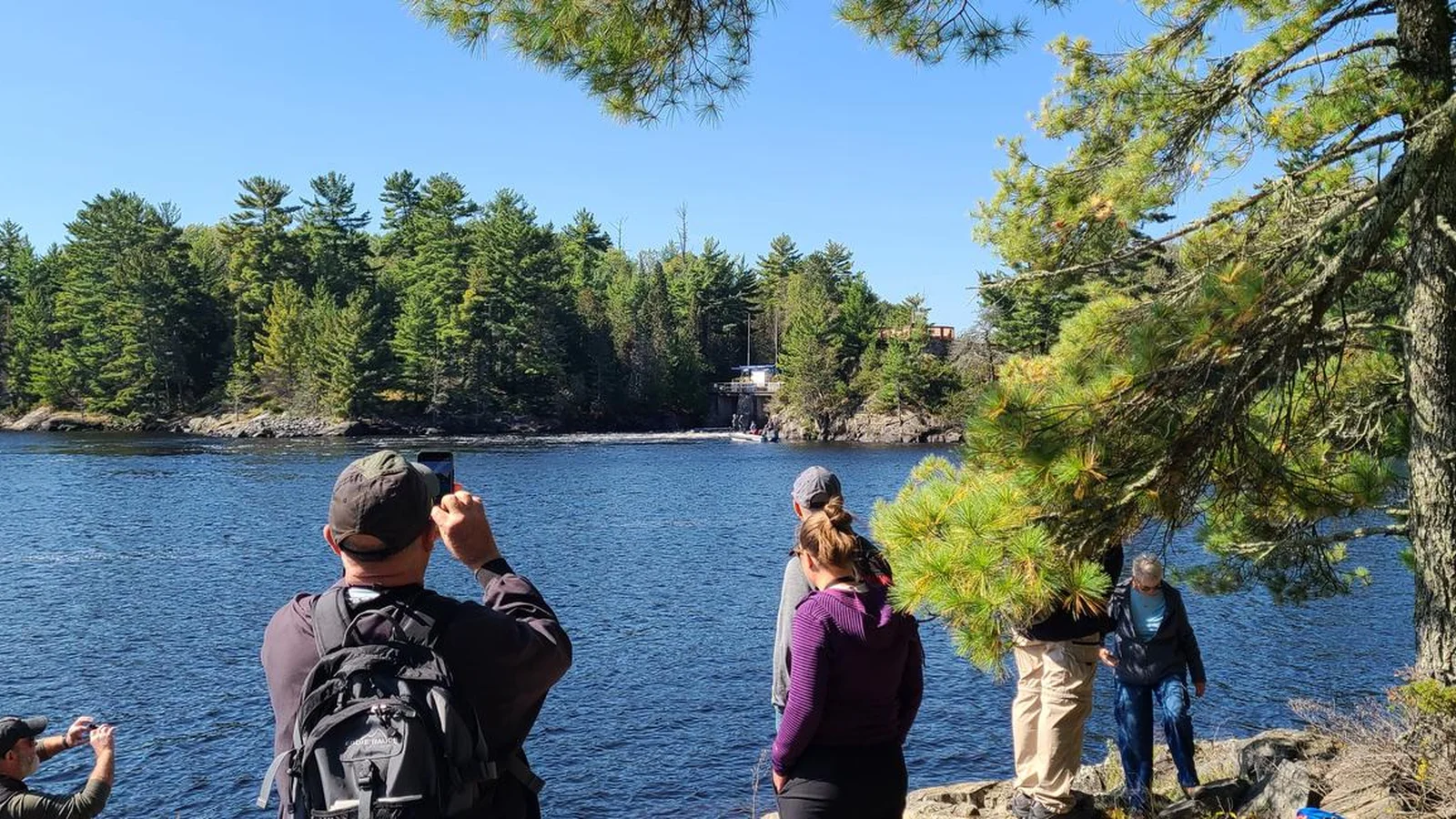 Visitors take photos of the Kettle Falls dam from a rocky, pine-covered shoreline