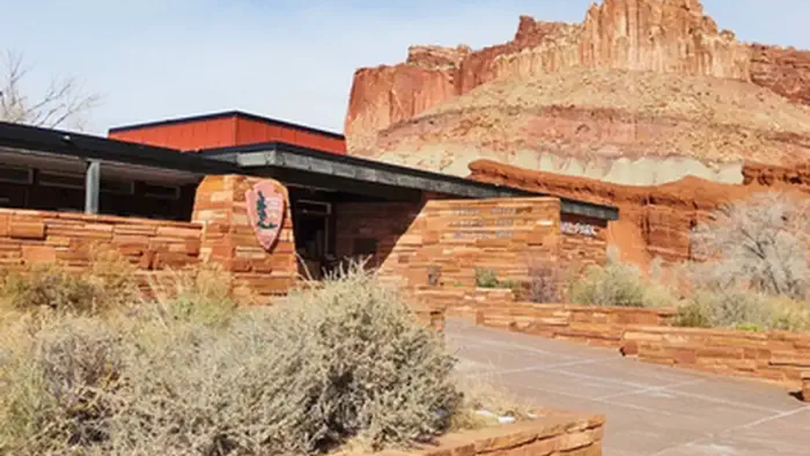 A brown stone building. The NPS Arrowhead is on the left side of the building. Desert vegetation is in front of the building. Orang-red cliffs rises above the building. They sky is cloudy with patches of blue.