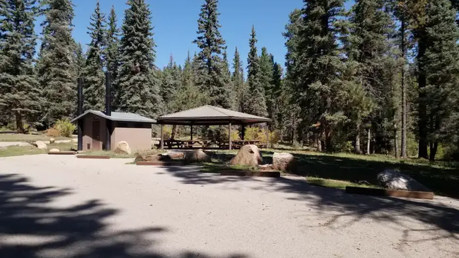 A parking area with a group shelter and vault toilet in a forest.