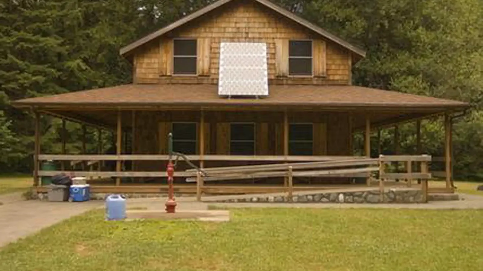 House with shingle siding,  solar panel on the roof of wrap around covered porch, hand pump well on the lawn, and ramp leading up to porch