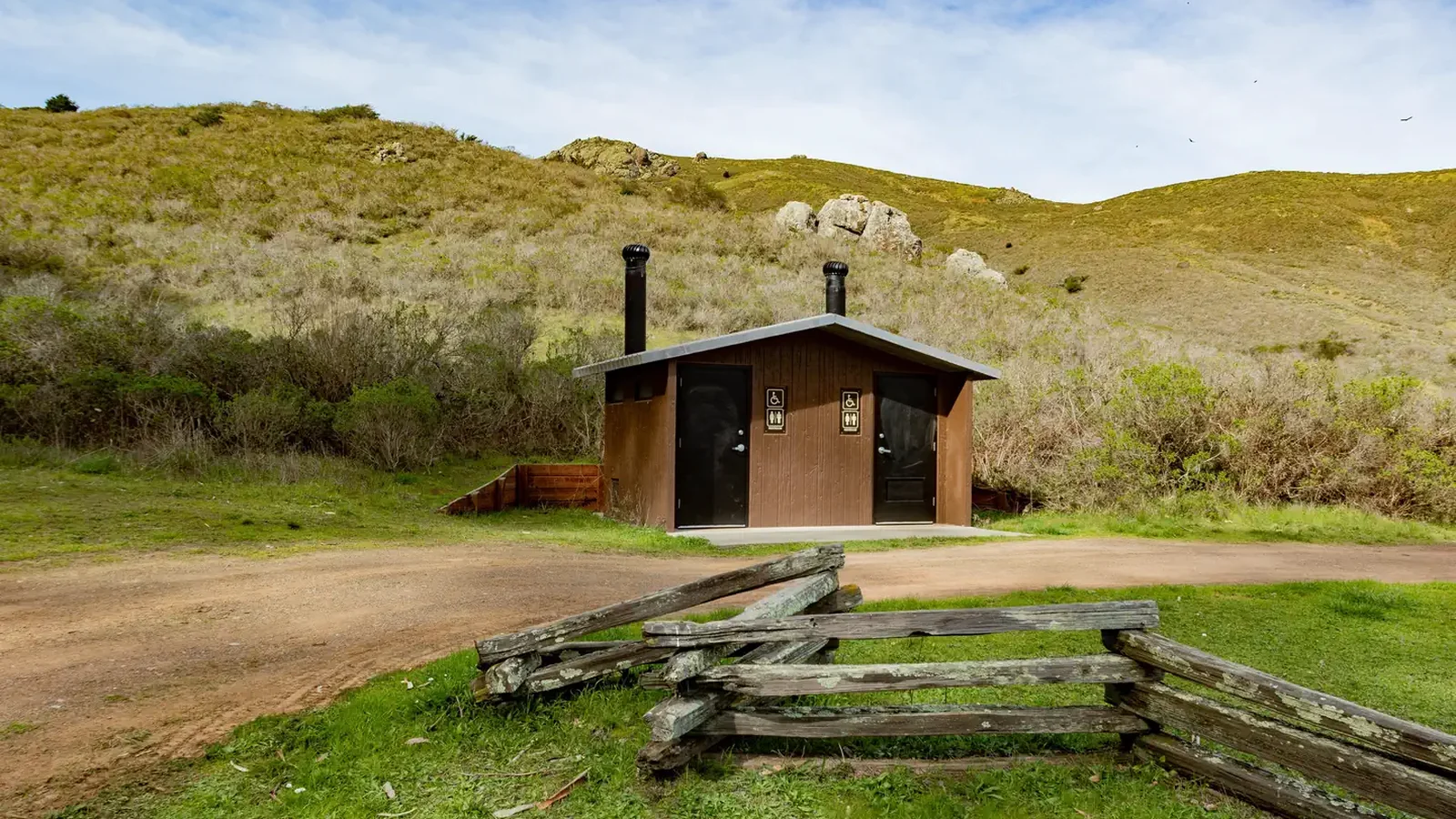 A pit toilet structure sits in front of along a dirt path. In the distance are shrub covered hills.