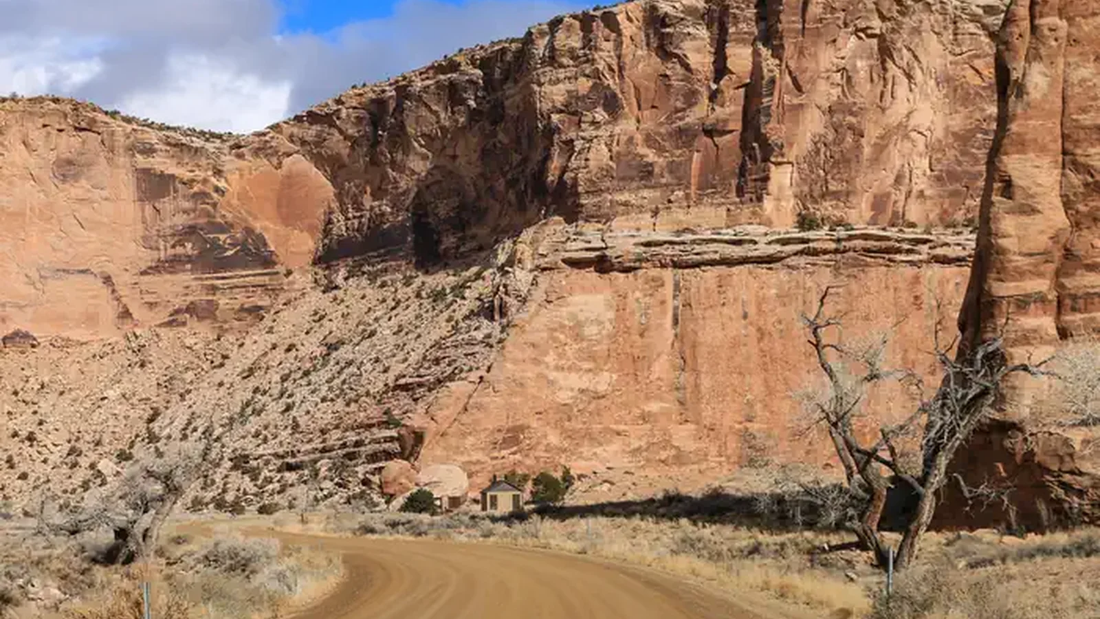 A dirt road cuts through a wide canyon of orange cliffs with a bathroom in the distance.