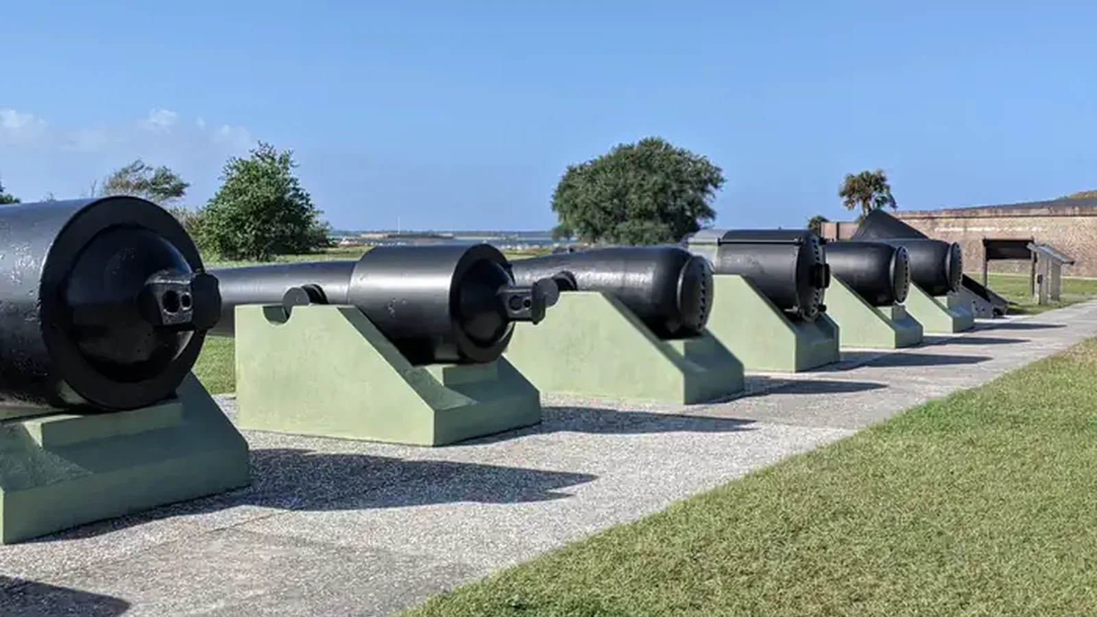 A blue sky appears over cannon row, an outdoor display of 7 black U.S. Civil War era cannon set on green cannon carriages.