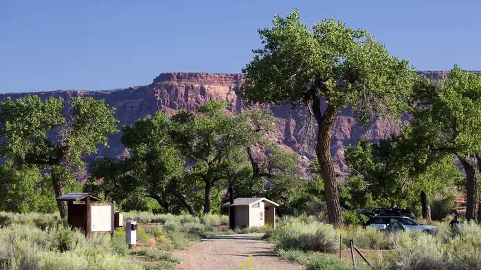 A registration kiosk and and vault toilet under Cottonwood trees with red rock cliffs in the background.