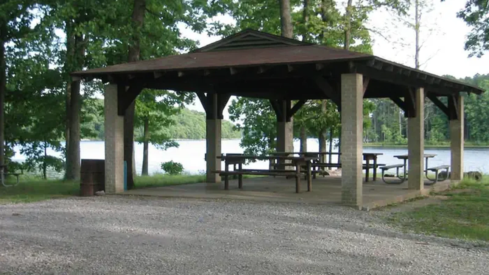 A pavilion with picnic tables, concrete floor and brick columns. The pavilion is next to a lake and shaded by trees. A gravel road connects to the pavilion.