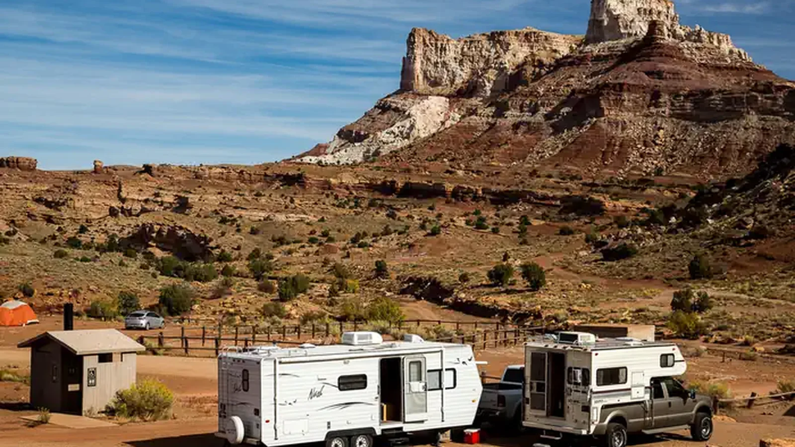 An RV and truck camper are parked in a campground below a large sandstone butte.