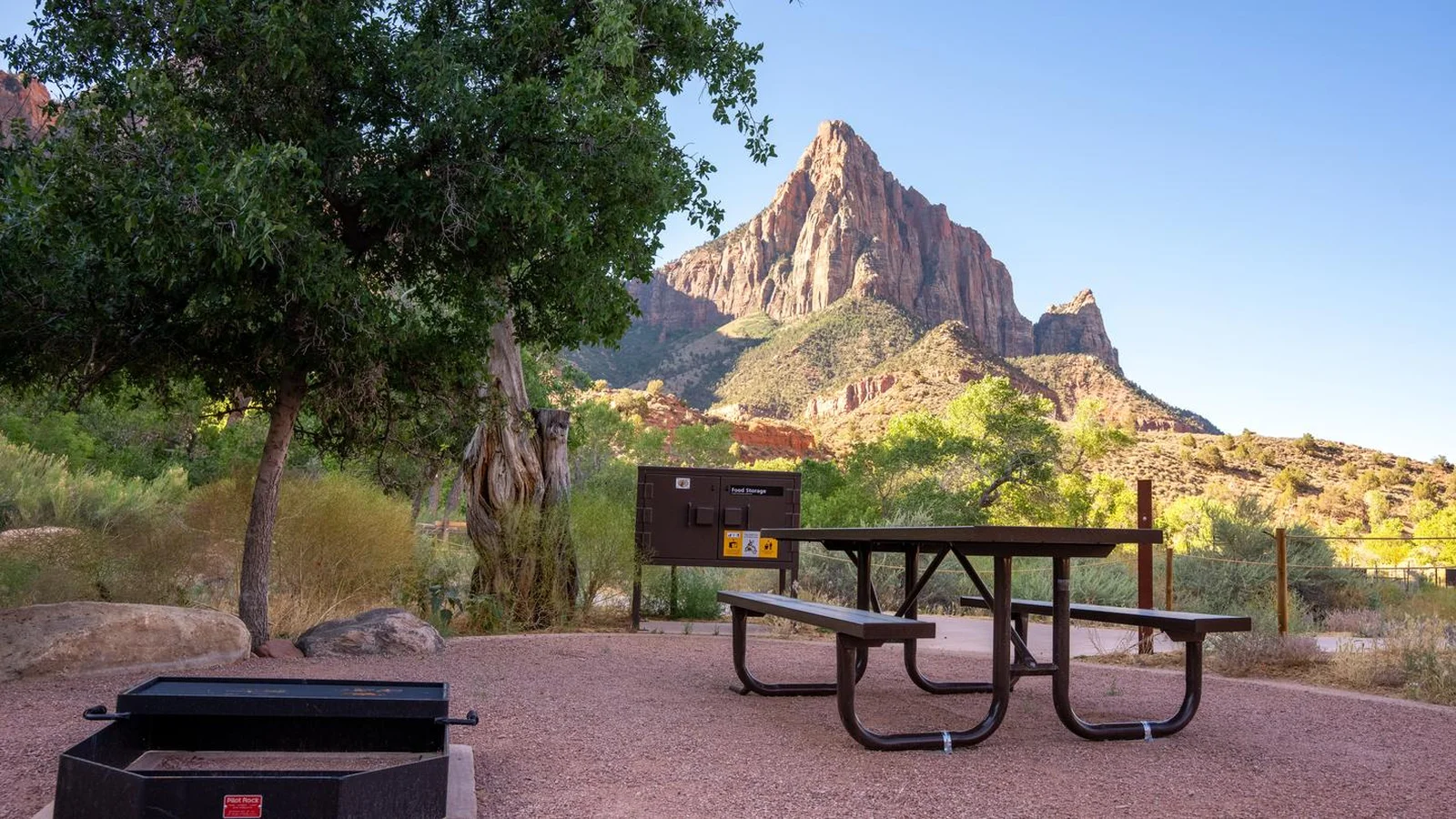 A campsite with a picnic table, fire ring, and food storage locker sits among low vegetation, with rugged sheer sandstone cliffs rising in the background.