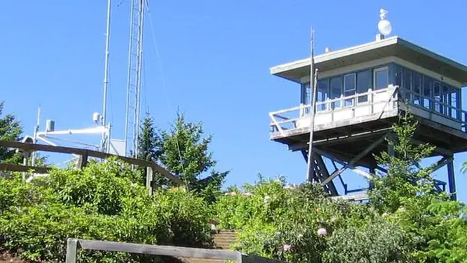 Lookout building on ridge with Forest Service i.d. sign, communications tower, green shrubs and conifer trees backed by blue sky.