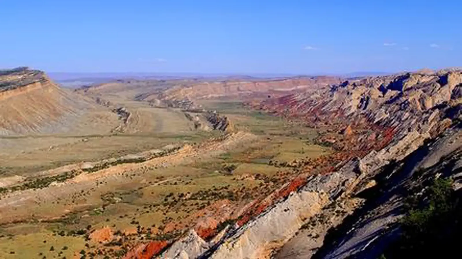 A green valley, a brown ridge cuts through it. Red and brown cliffs slope up on the right side of image. The sky is a bright, cloudless blue.