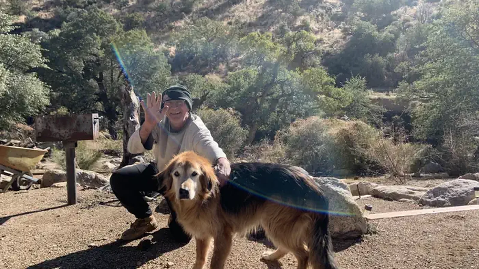 A man squatting down next to his dog, the man is smiling and waving at the camera.