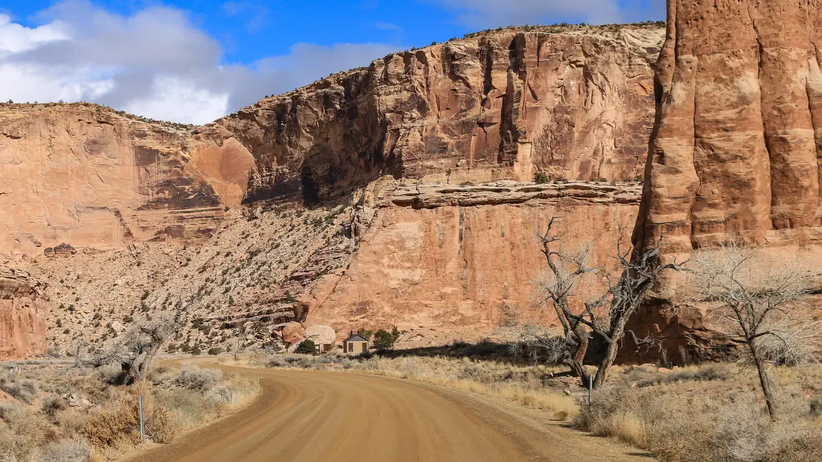 A dirt road cuts through a wide canyon of orange cliffs with a bathroom in the distance.