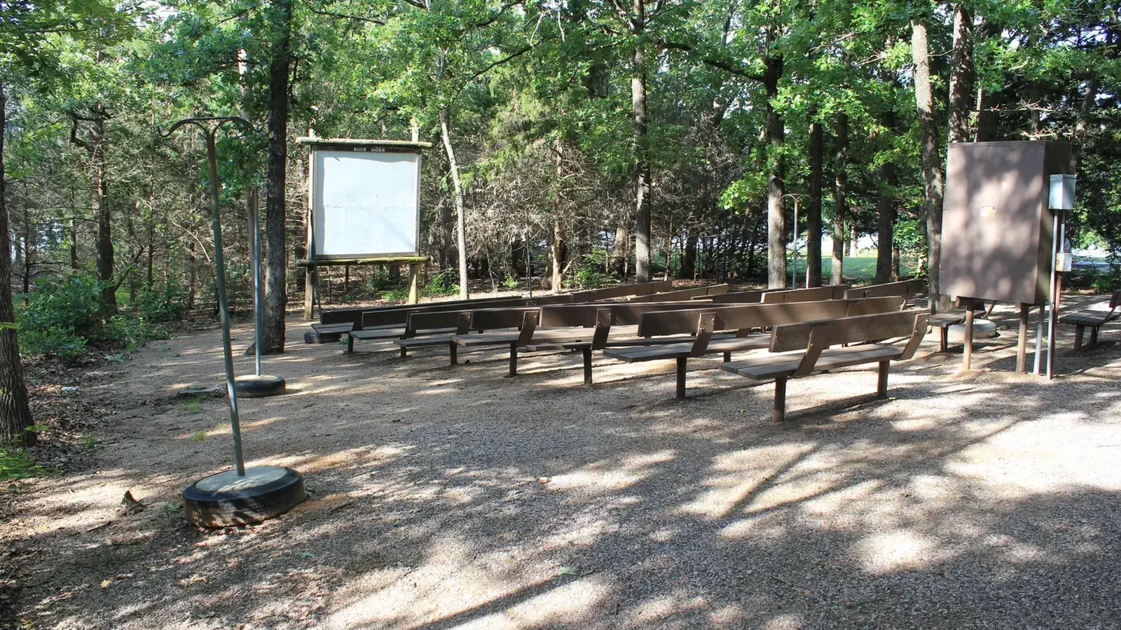 surrounded by trees, an ampitheater with wooden benches faces away from the viewer. A white projector board is in front of the benches.