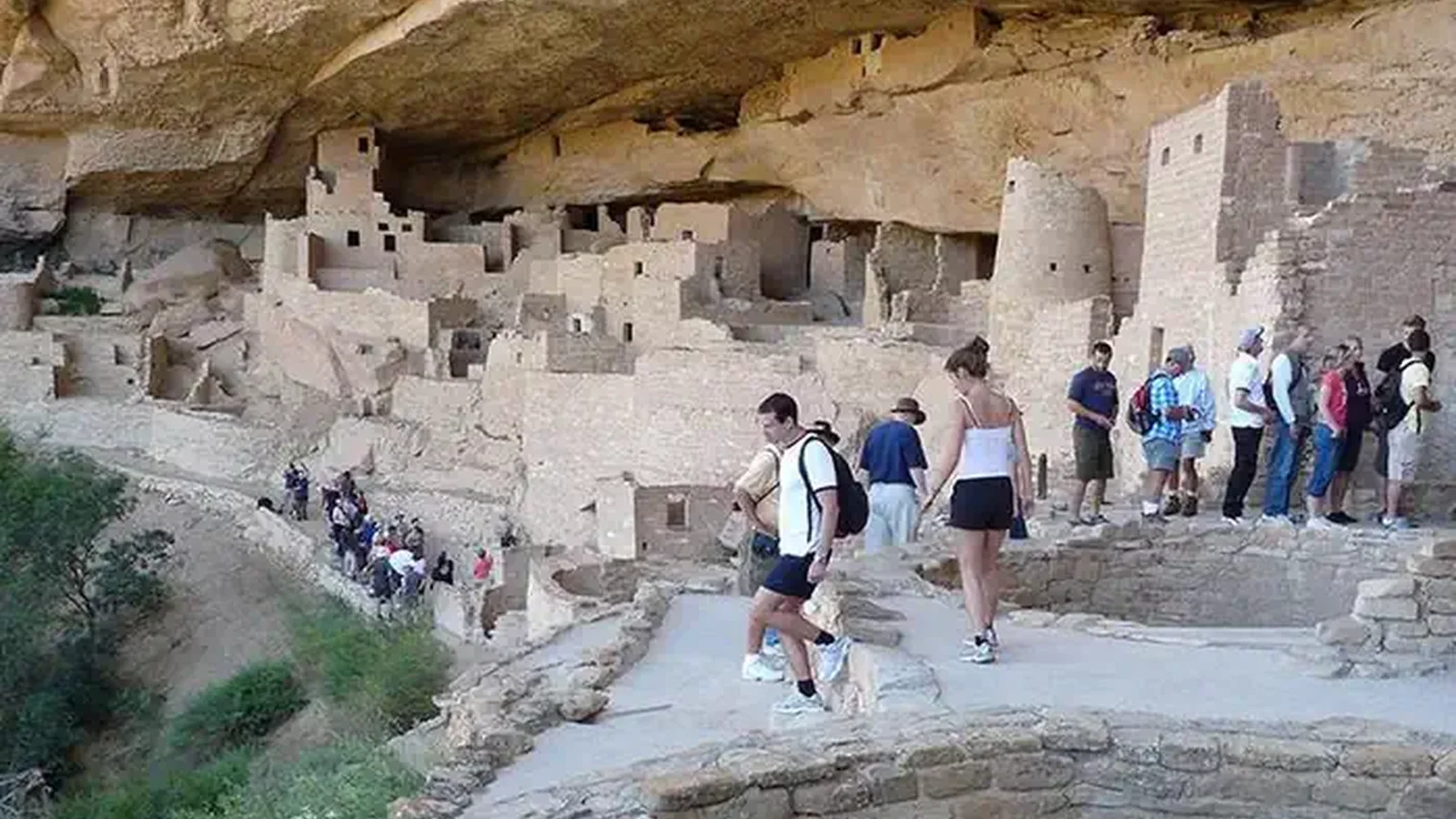 A variety of people walking around a large, ancient, stone-masonry village in a cliff alcove, with stone above and to the rear of the buildings and trees and vegetation on the slope to the left..